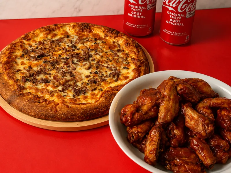 A pizza with a bowl of saucy chicken wings and two cans of Coca-Cola
